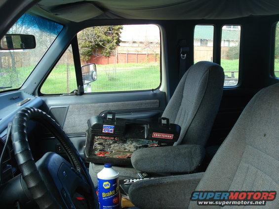 Interior of a truck cab, showing the steering wheel, seats, and a toolbox. A bottle of brake fluid is on the floor
