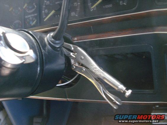 Close-up of a vehicle's dashboard with vice grips clamped onto the steering column. Gauges are visible behind the steering wheel