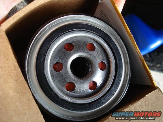 Oil filter with red filter media visible through six holes, sitting in a cardboard box
