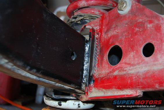 Close-up of a red metal bracket with two holes, welded to a black metal beam. A red coil spring is visible