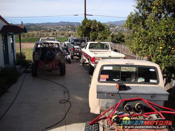 Several trucks and off-road vehicles parked in a driveway, with a house and hills in the background