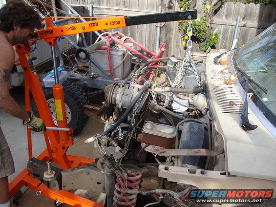 Engine hoist lifting a V8 engine from a tan 1980s SUV with exposed wiring and a rusty brake master cylinder