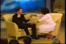 Man in black shirt and woman in pink outfit on yellow couch, audience in foreground