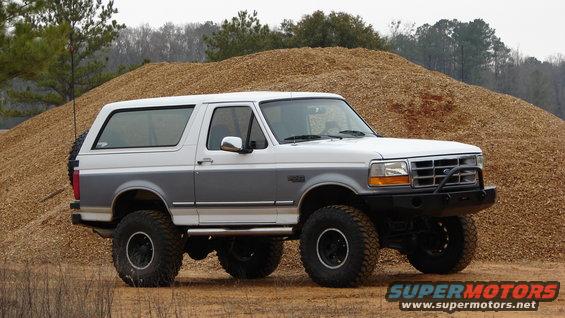 White and gray Ford Bronco with aftermarket bumper and large tires parked in front of a wood chip pile