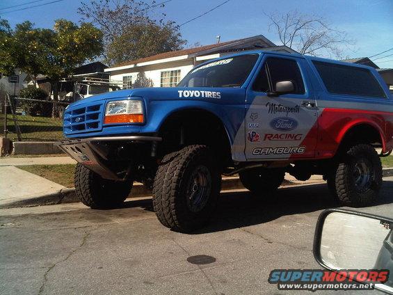 Blue and silver Ford Bronco with racing decals, lifted with off-road tires, parked on a street
