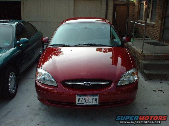 Front view of a red Ford sedan parked next to a dark green car. License plate V75-LYM is visible