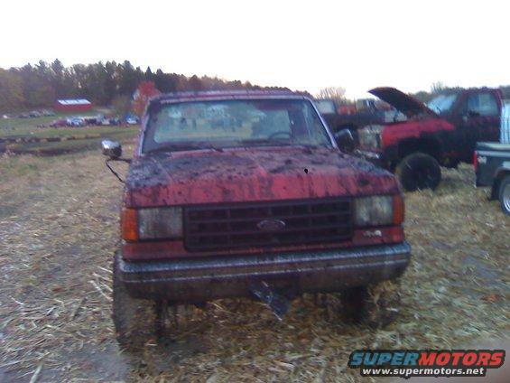 Mud-covered red pickup truck, front view. Another red truck with hood up in background