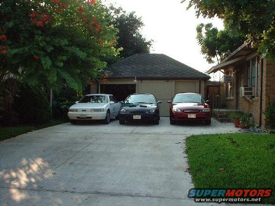 Three cars parked in front of a garage: a white sedan, a black sedan, and a red sedan