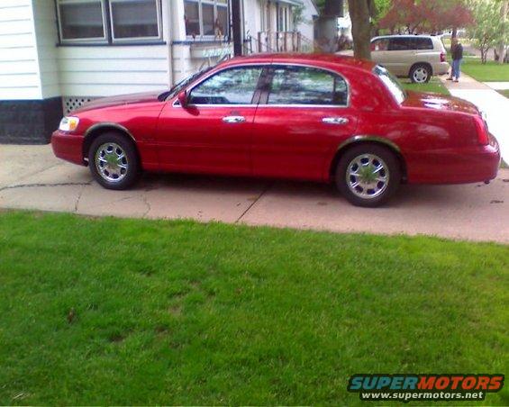 Red 1999 Lincoln Town Car parked on a driveway next to a house. Another SUV is visible in the background