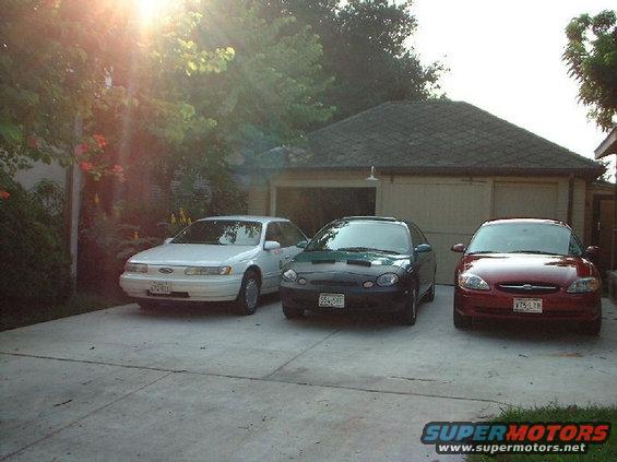 Three sedans parked in a driveway. The white car on the left is a Ford Taurus. The dark green car in the middle and the red car on the right are also Ford Tauruses