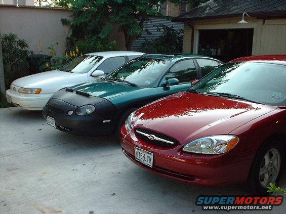 Three Ford Taurus sedans parked in a driveway. The front car is red, the middle is green with a car cover, and the rear is white