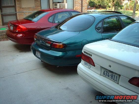 Three sedans parked in a lot. The closest is white, the middle is teal, and the furthest is red. The teal car has 'TAURUS' badging