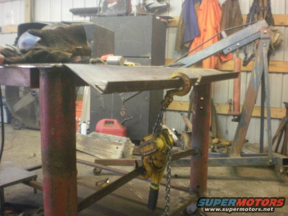 Engine hoist and chain attached to a metal workbench in a garage. Tools and equipment are visible in the background