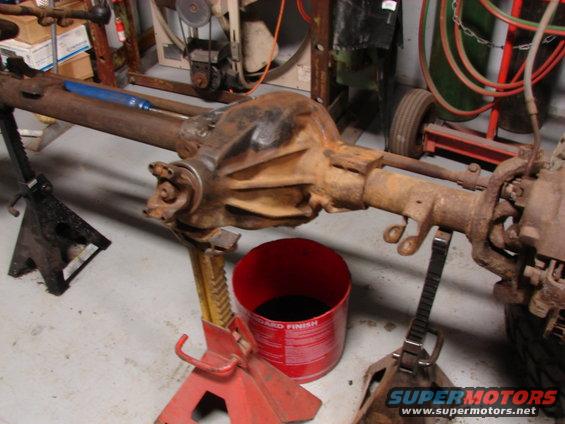 Rusty rear axle assembly on jack stands with a red bucket of fluid