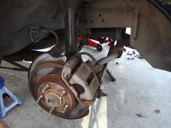 Close-up of a car's rear brake rotor, caliper, and suspension components. A blue jack stand is visible