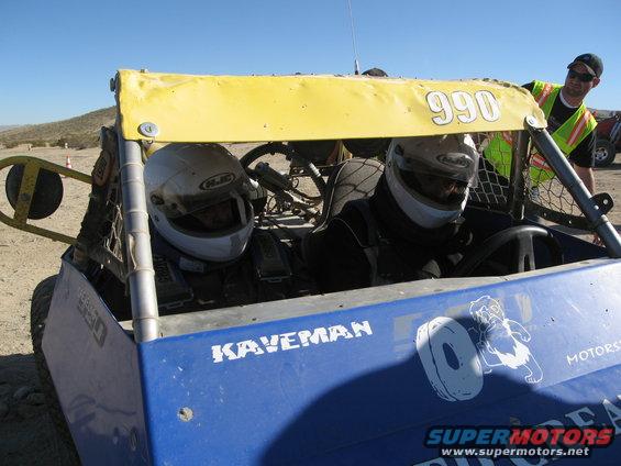 Blue off-road buggy interior with two helmeted drivers and a yellow sunshade labeled 990