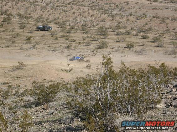 Two off-road vehicles driving in a dusty desert landscape with sparse vegetation