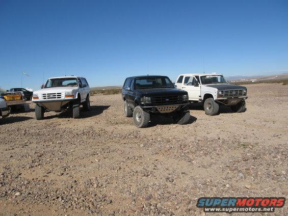 Three Ford Bronco SUVs parked on a gravelly surface under a clear blue sky. The front vehicle is white, the middle is black, and the rear is white with a black hood