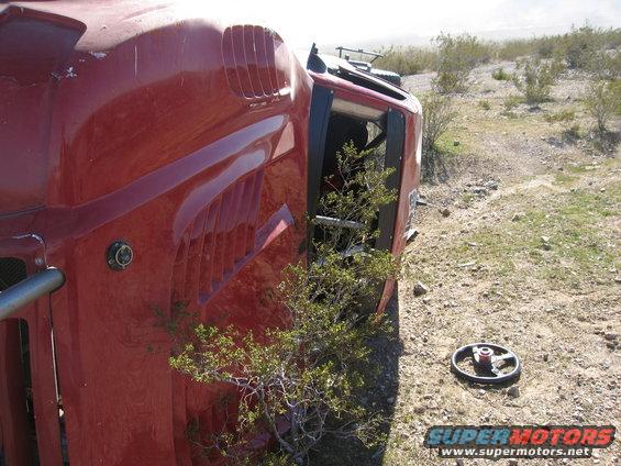 Red vehicle flipped on its side in a desert landscape. A small bush is growing through the open door