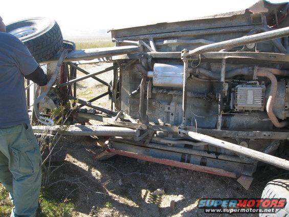 Overturned vehicle chassis with exposed undercarriage, exhaust, and suspension components. A spare tire is visible