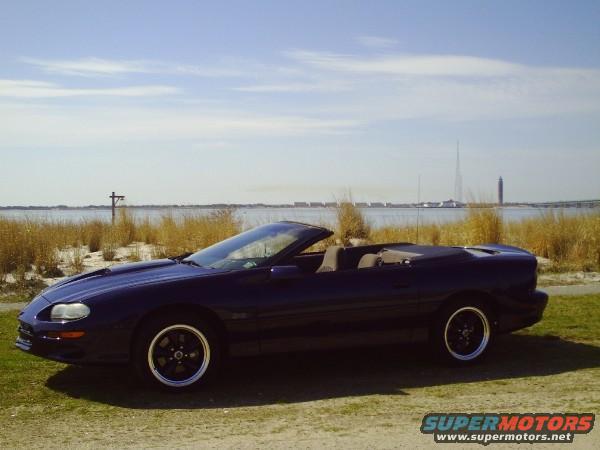 Dark blue 2001 Chevrolet Camaro convertible parked by a body of water with dry grass and a distant tower