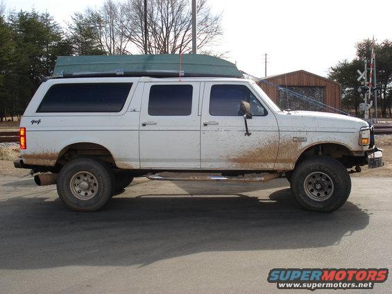 White extended cab pickup truck with a green boat on its roof rack, parked on a road with mud splatters on the side
