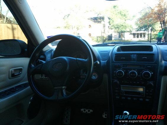 Car interior with black steering wheel, tan door panel, and center console with radio and climate controls. Supermotors.net logo visible
