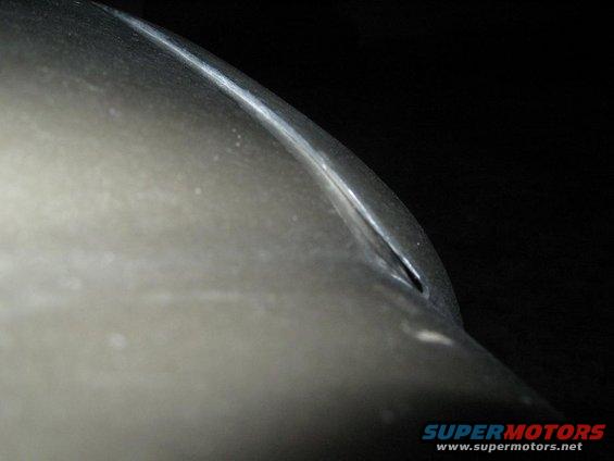 Close-up of a silver car body panel with a sharp crease and a small opening, against a black background