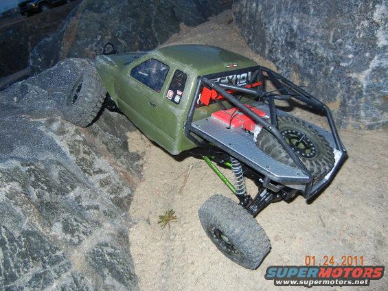 Green RC rock crawler with roll cage and spare tire on rocky terrain