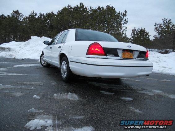 White Ford Crown Victoria sedan parked on wet asphalt with snow piles in background. License plate reads FGV-7260