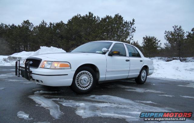 White police car with push bumper parked on a wet, icy road with snow and trees in the background