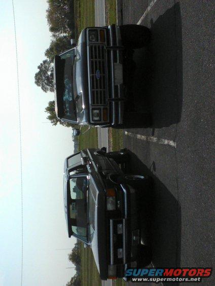 Two dark-colored Ford Broncos parked side-by-side, front view. The front Bronco has a chrome grille and bumper