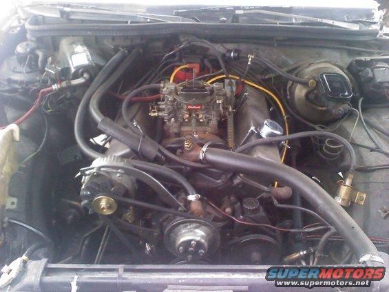 Engine bay showing V8 engine with carburetor, alternator, and various hoses and wires