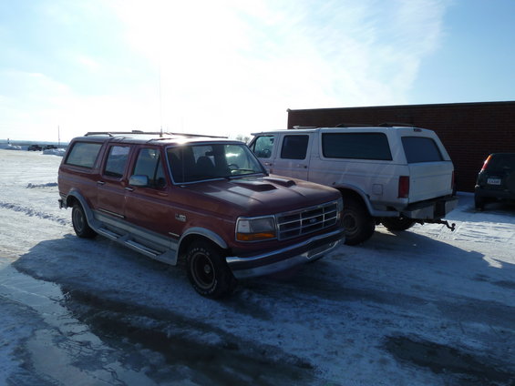 Red extended cab pickup truck with camper shell, 3/4 front view. Another white SUV is behind it