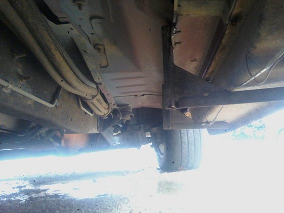 Underside of a vehicle showing frame, fuel lines, and a tire. The vehicle appears aged