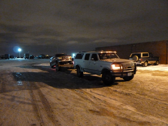 White Ford SUV towing a dark pickup truck at night in a snowy lot