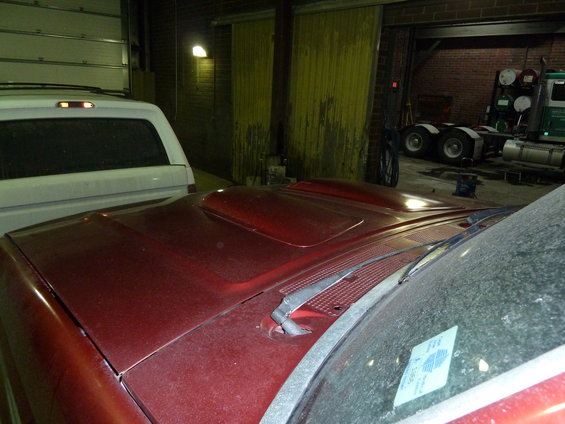 Red pickup truck hood with a hood scoop, parked next to a white SUV. A semi-truck is visible in the background