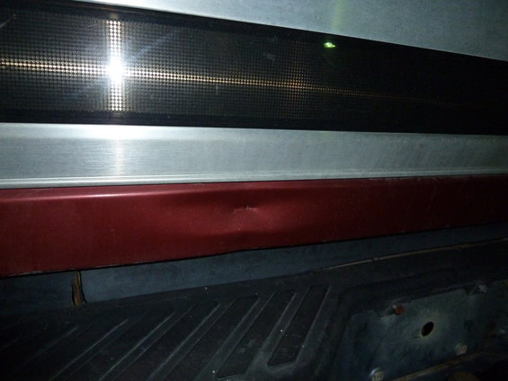 Dent on red body panel above black textured surface, possibly a truck bed or bumper area