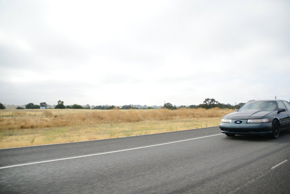 dark sedan driving on a highway with dry grass fields and trees in the background