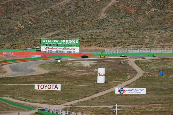 Race track with a dark sedan, yellow sports car, and other vehicles on the track under a cloudy sky