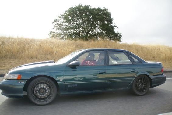Green 1995 Ford Taurus sedan driving on a road with two people inside