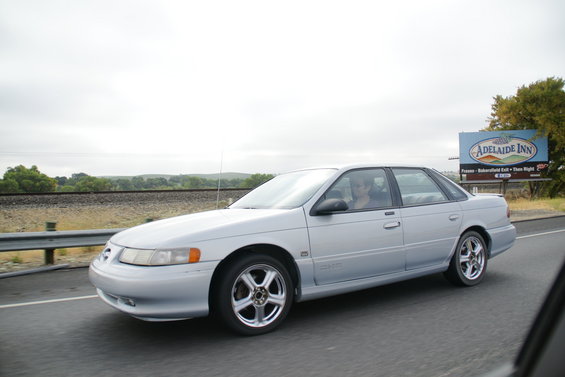 Silver sedan driving on highway, side view. Looks like a 1996 Ford Taurus