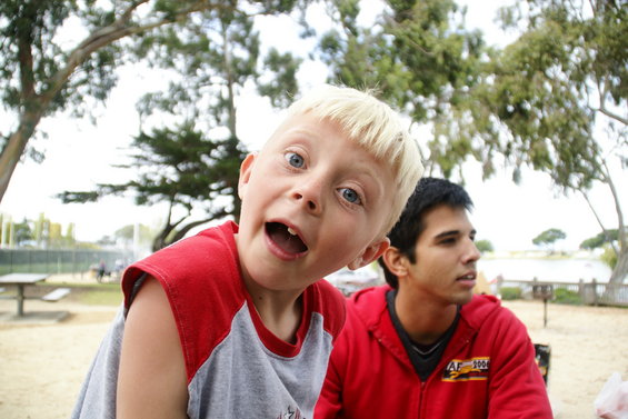 Kid with blond hair and blue eyes makes a funny face in front of a guy in a red hoodie