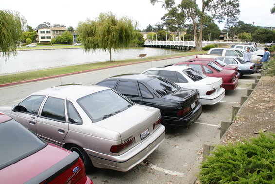 Several sedans parked in a lot near a lake. The closest car is a tan sedan, followed by a black sedan, a white sedan, and a red sedan