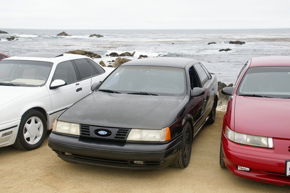 Black 1995 Ford Taurus parked between a white sedan and a red sedan near the ocean