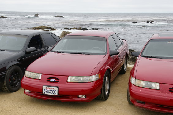Red sedan with California plates 5ZDS335 parked next to a black sedan and another red sedan by the ocean