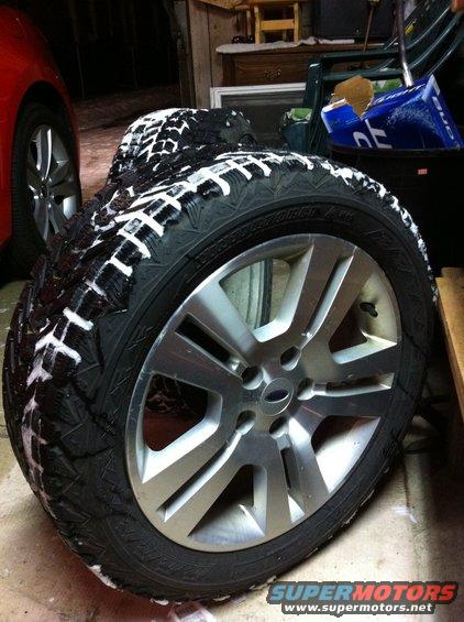 Close-up of a winter tire with white markings on a silver alloy wheel, with a red car visible in the background