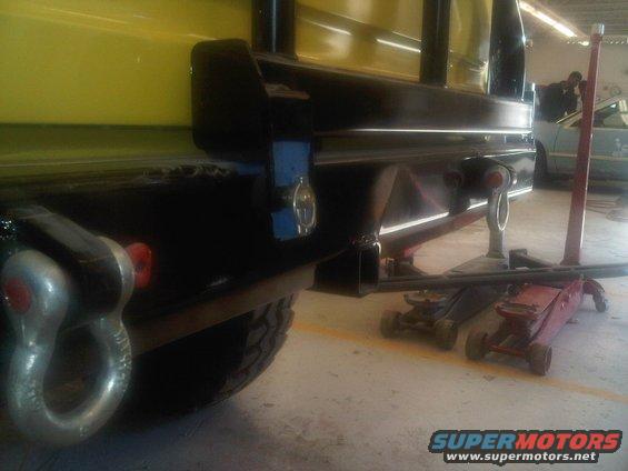 Close-up of a heavy-duty tow hitch and shackle on a vehicle's rear bumper. Two red floor jacks are visible underneath