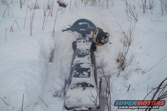 Snowmobile partially buried in snow, with dry grass and branches around it. A helmet rests on the seat