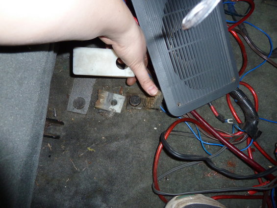 Car interior floor with exposed wiring, mounting brackets, and a gray vent cover being held by a hand